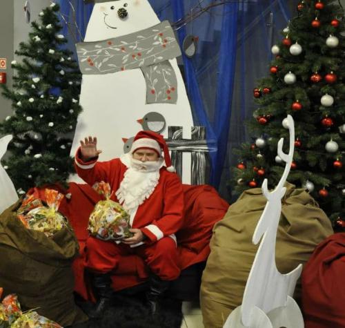 Mikołaj z podniesioną ręką na tle bałwana i choinek. Przed mikołajem prezenty i biały renifer.