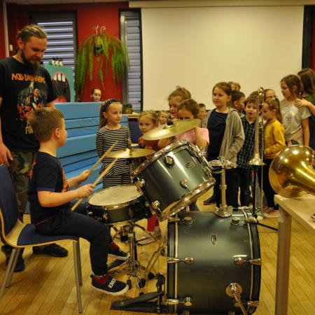 Chłopiec gra na perkusji, za nim instruktor i wiele dzieci w kolejce