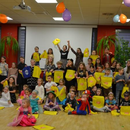 Grupa dzieci pozuje do zdjęcia. Wiele z nich trzyma w ręku żółte woreczki, nad nimi balony