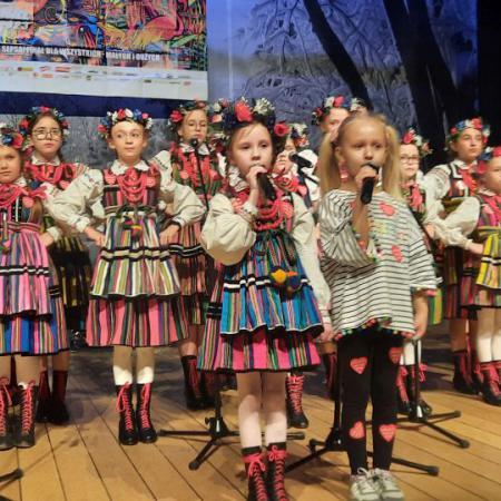 Dziewczynki śpiewają przy mikrofonach , za nimi grupa dzieci w ludowych strojach