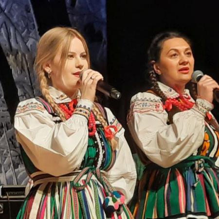 Dwie kobiety w ludowych strojach śpiewają przy mikrofonach na scenie w RCK