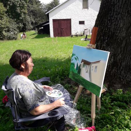 Kobieta siedzi pod Drzewem i maluje obraz przedstawiający Dworek 