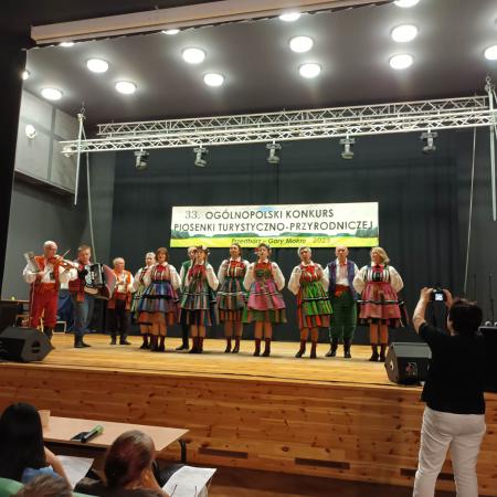 Grupa osób w ludowych strojach śpiewa na scenie podczas Konkursu Piosenki Turystyczno-przyrodniczej
