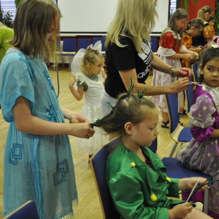 Dzieci i dorośli w przebraniach robią fryzury przed lustrem innym dzieciom