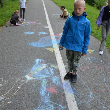 Na pierwszym planie stoi chłopiec , za nim dzieci rysują kredami 