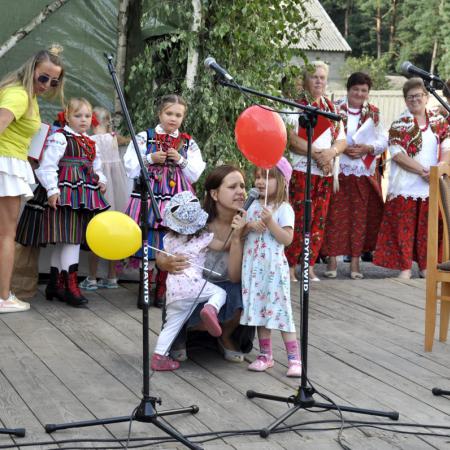 Dwie małe dziewczynki z balonikami na pierwszym planie, jedna z nich śpiewa do mikrofonu za nimi inne dzieci i kobiety