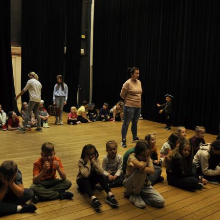 Dzieci siedzą w dwóch rzędach na scenie , obok nich stoja instruktorki zajęć.