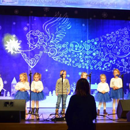 Dzieci w wieku przedszkolnym spiewają na scenie, w tle Mikołaj.