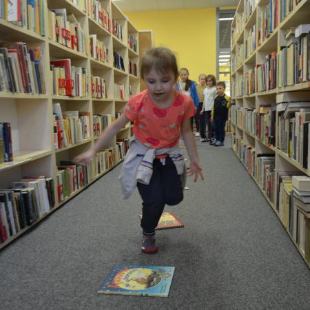 Dziewczynka skacze przez książki ułożone na dywanie w bibliotece