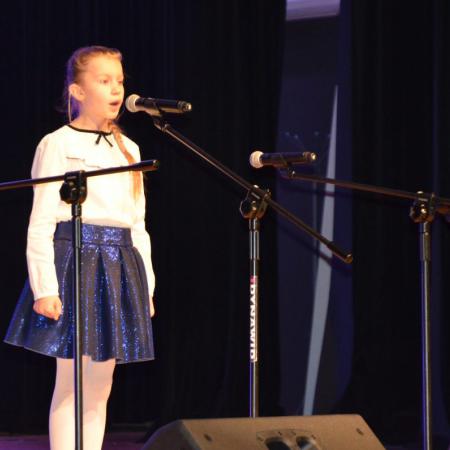 Dziewczynka w niebieskiej spódniczce przy mikrofonie na scenie.