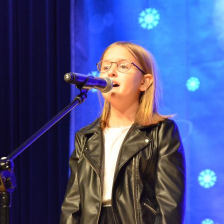Dziewczynka w okularach i czarnej kurtce, śpiewająca na scenie