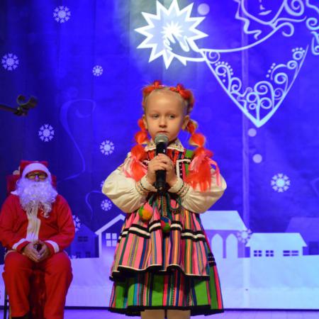 Dziewczynka w wieku przedszkolnym w stroju ludowym ,trzymająca mikrofon w ręce , w tle Mikołaj.