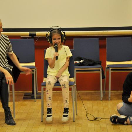 Dziewczynka z mikrofonem i słuchawkach siedzi na krzesełku i śpiewa piosenkę, obok siedzą instruktorki zajęć.