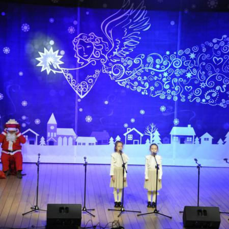 Dziewczynki w wieku przedszkolnym śpiewają na scenie, w tle Mikołaj