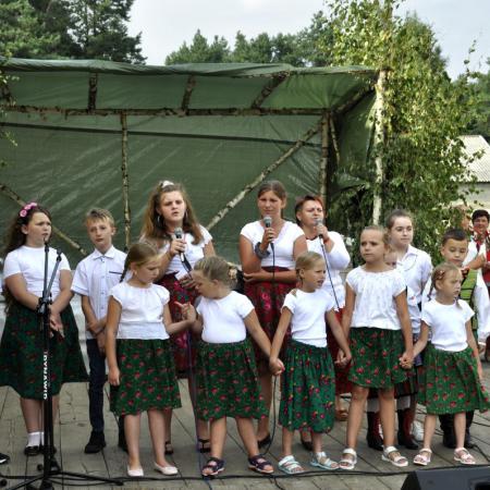 Grupa dzieci śpiewa do mikrofonów. Dziewczynki w zielonych i czerwonych spódnicach, chłopcy w białych koszulach