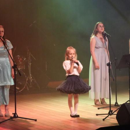 Mała dziewczynka śpiewa przy mikrofonie na scenie , za nią chórek starszych dziewczyn