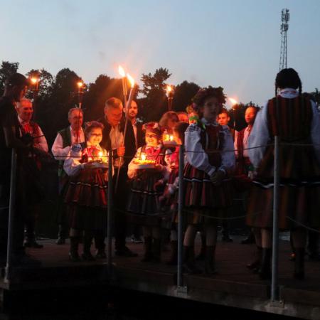 Mężczyźni z pochodniami, dziewczyny i chłopaki z w strojach ludowych stoją na pomoście nad zalewem