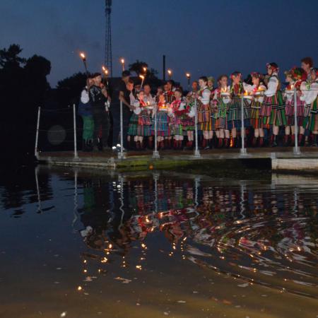 Grupa osób w strojach ludowych z pochodniami śpiewa na molo nad zalewem