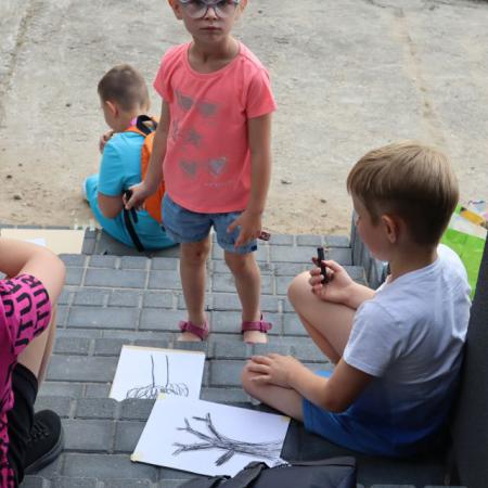 Grupa dzieci siedzi i maluje obrazy, na środku stoi dziewczynka
