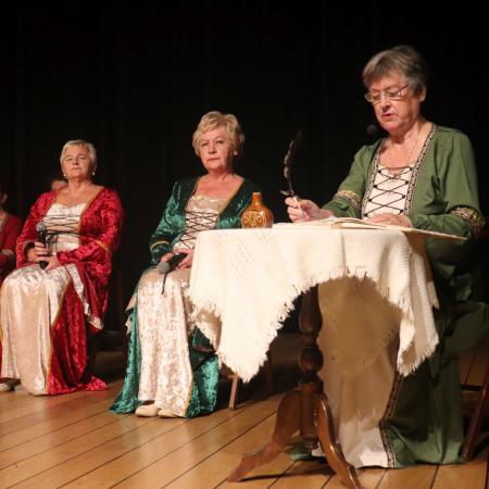 Kobieta w zielonej sukience siedzi przy stoliku i trzyma pióro, obok niej siedzą dwie kobiety 