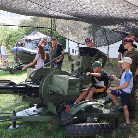 Dzieci siedzą na maszynach wojskowych obok nich stoją żołnierze w mundurach