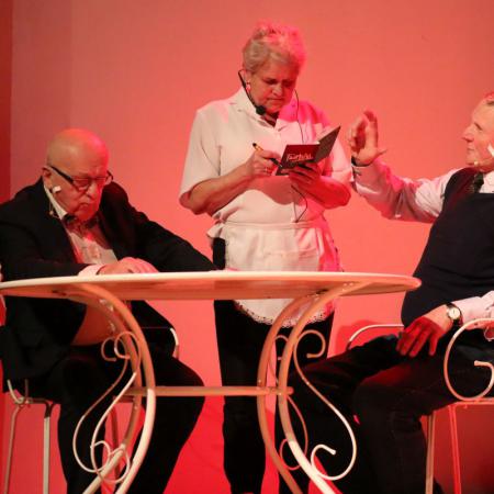 Dwóch mężczyzn siedzących przy stole i kobieta grająca kelnerkę występują na scenie podczas sztuki pt. "Kluczyk" 