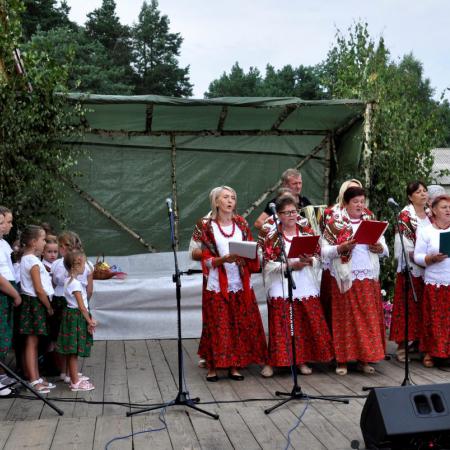 Kobiety w czerwonych sukienkach śpiewają do mikrofonów, obok stoją dzieci