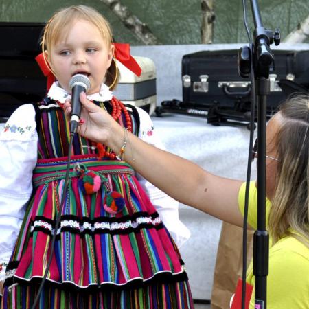 Malutka dziewczynka w kolorowym stroju śpiewa, starsza pani podtrzymuje jej mikrofon