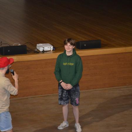 Mężczyzna w czerwonej czapce , z mikrofonem stoi zwrócony w stronę chłopaka w zielonej bluzie.