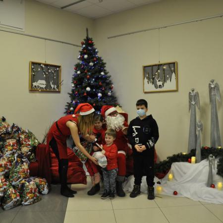 Mikołaj trzyma dziewczynkę na kolanach , Sieżynka wręcza prezent małemu chłopcu obok nich stoi chłopiec z prezentem
