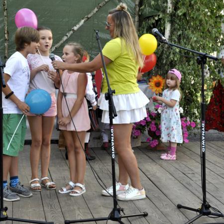 Troje dzieci z balonikami śpiewa do mikrofonu, kobieta przytrzymuje im mikrofon